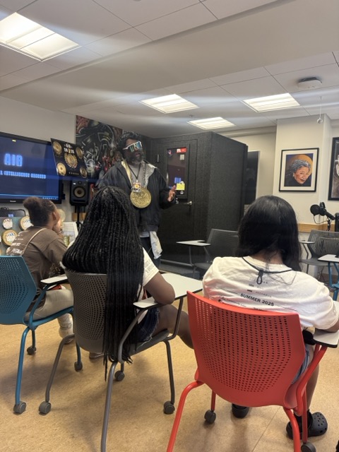 A man with a wig and chain speaking to students in a classroom